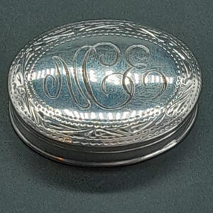 May include: Oval silver trinket box with engraved floral border and monogram. The lid features a raised monogram in a stylized font. The box is made of a shiny, reflective metal.