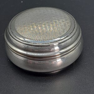 Op de afbeelding: Een ronde zilveren sieradendoos met een getextureerd deksel. Het deksel heeft een verhoogde rand en een glad, glanzend oppervlak.