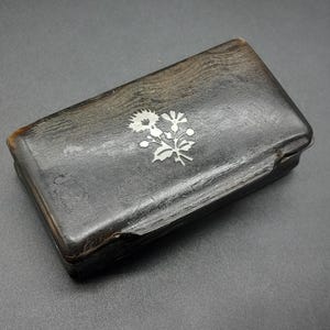 以下が含まれることがあります： 長方形の濃い茶色の箱で、蓋には銀色の花の模様があります。箱は質感のある表面で、革または同様の素材で作られているようです。箱の端はわずかに摩耗しています。