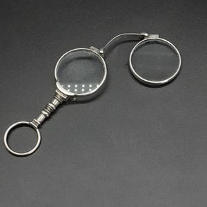 Op de afbeelding: Een zilverkleurige vouwloep met twee lenzen. De loep heeft een handvat met een ring aan het einde.