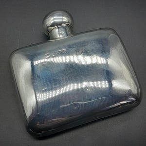 Pode incluir: Uma garrafa retangular prateada com um topo arredondado e uma pequena rolha esférica. A garrafa tem uma superfície refletora e a palavra "3000" está estampada na frente. O item é provavelmente feito de metal.