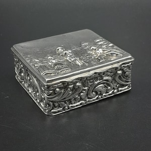 Puede incluir: Una caja rectangular de metal plateado con una tapa en relieve que muestra una escena de figuras en un paisaje. Los lados de la caja están decorados con intrincados patrones elevados de volutas florales y hojas.