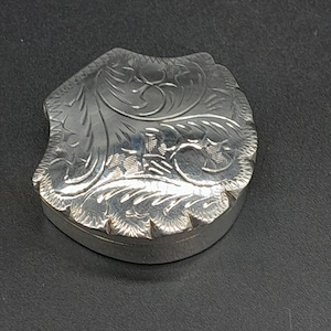 May include: A small, silver-colored metal pill box with a shield-like shape. The lid features an intricate, engraved floral pattern with swirling leaves and fine line details. The sides of the box are smooth and polished, reflecting light.