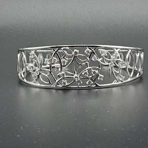 Könnte beinhalten: Ein silbernes Armreif-Armband mit einem zarten Blumenmuster und klaren Steinen.