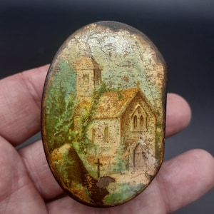 May include: Oval-shaped, antique metal piece with a painted church scene. The artwork depicts a stone church with a steeple, surrounded by trees and a cross. The piece has a distressed, aged appearance.