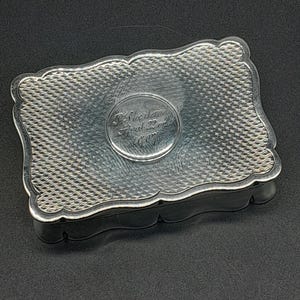 Puede incluir: Una caja rectangular de plata con borde festoneado y un patrón de sombreado cruzado texturizado en la tapa. Un panel central circular liso presenta el texto grabado: P. Sterling, April 22nd, 1907. El metal tiene un acabado pulido y reflectante.