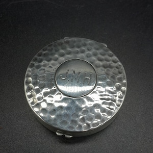 Puede incluir: Un estuche compacto redondo de color plateado con una textura martillada. El centro presenta un área circular más pequeña con las iniciales "J.M.H." en relieve. El estuche compacto tiene un cierre de bisagra y un borde ligeramente elevado.