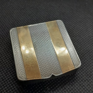 May include: A silver and gold compact with a textured design. The compact has three gold stripes on a silver background.