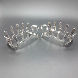 Könnte beinhalten: Zwei silberfarbene Toastständer im Art-Deco-Stil. Jeder Ständer hat eine Reihe vertikaler Stäbe, die Fingern ähneln und mit einer Basis verbunden sind. Die Ständer sind für mehrere Toastscheiben konzipiert. Der Hintergrund ist einfarbig dunkelgrau.