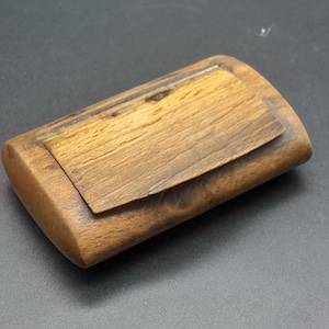 May include: A rectangular wooden box with a hinged lid. The box is made of light brown wood with a visible grain pattern. The lid is slightly darker than the body. The box is closed and rests on a dark surface.