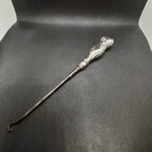 Puede incluir: Un calzador de plata con un mango retorcido y un gancho en el extremo.