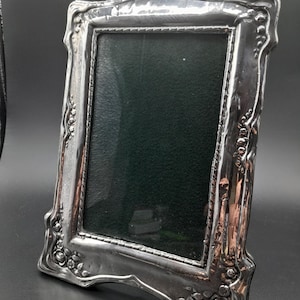 May include: A silver picture frame with an elaborate floral pattern. The frame has a rectangular aperture with a dark green backing. The frame is angled, highlighting its decorative details and reflective surface.