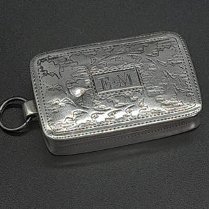 May include: A rectangular silver-toned metal box featuring an intricate embossed pattern across the lid and a central rectangular plaque engraved with the letters EM. The box includes a small circular metal ring attached to one side for hanging.
