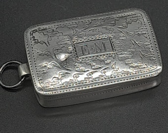 May include: A rectangular silver-toned metal box featuring an intricate embossed pattern across the lid and a central rectangular plaque engraved with the letters EM. The box includes a small circular metal ring attached to one side for hanging.