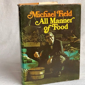 Puede incluir: Libro de cocina vintage con el título "All Manner of Food" de Michael Field. La portada presenta a un hombre con gafas sosteniendo alimentos en un mercado. El título está en escritura naranja y amarilla sobre un fondo verde oscuro.