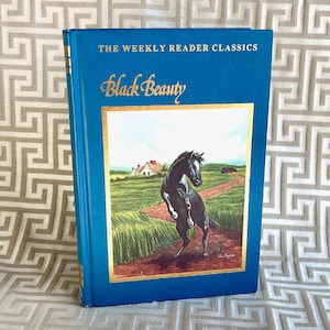 May include: A teal-coloured hardcover book titled "Black Beauty" from "The Weekly Reader Classics". The cover illustration shows a black horse rearing up on a path, with a house in the background. Gold accents frame the illustration.
