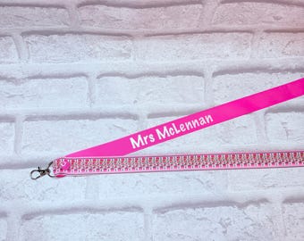 May include: A bright pink lanyard featuring the white text Mrs McLennan printed on the fabric. The strap includes a decorative trim with a repeating pattern of small silver metallic accents and a silver-toned metal lobster clasp at the end.