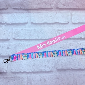 May include: A dual-layered lanyard featuring a solid pink fabric strap with the white text Mrs Hamilton printed on it, layered over a blue fabric strap decorated with a repeating pattern of colourful books and potted flowers, finished with a metal clasp.