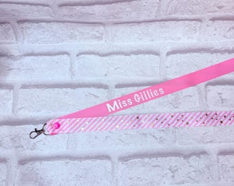 May include: A pink lanyard featuring the white text Miss Gillies and a second strap layer with a white and pink diagonal striped pattern accented by small gold stars, finished with a silver metal lobster clasp and a pink plastic snap button.
