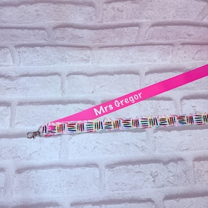 Peut inclure: Un cordon tour de cou avec une section rose vif portant le texte blanc Mrs Gregor et une section blanche ornée d'illustrations de crayons colorés. Le cordon comprend un petit bouton rose et un fermoir mousqueton en métal argenté.