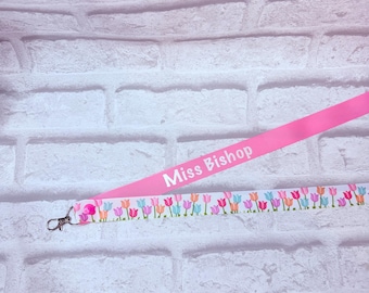 May include: A pink lanyard featuring the white text Miss Bishop and a white section decorated with a repeating pattern of colorful tulips in shades of pink, orange, and blue, finished with a silver-toned metal lobster clasp attachment.