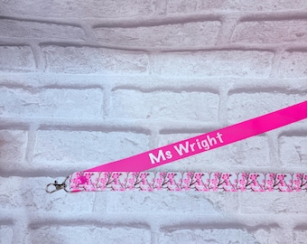 May include: A pink lanyard with the text "Ms Wright" printed in white. The lower portion of the lanyard features a floral pattern with pink blossoms and black branches. A silver-colored clasp is attached.