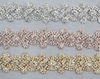 May include: Three decorative rhinestone trims in silver, rose gold, and gold. Each trim features a repeating floral pattern with clear, sparkling rhinestones. The trims are displayed on a light gray background, showcasing their intricate designs.