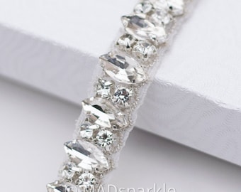 May include: A close-up of a silver and clear crystal bridal sash with a row of marquise-shaped crystals and smaller round crystals. The sash is on a white background.