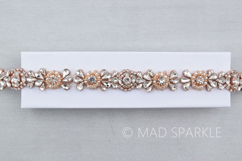 May include: A rose gold beaded and rhinestone trim with clear and pearlescent stones. The trim is on a white background.