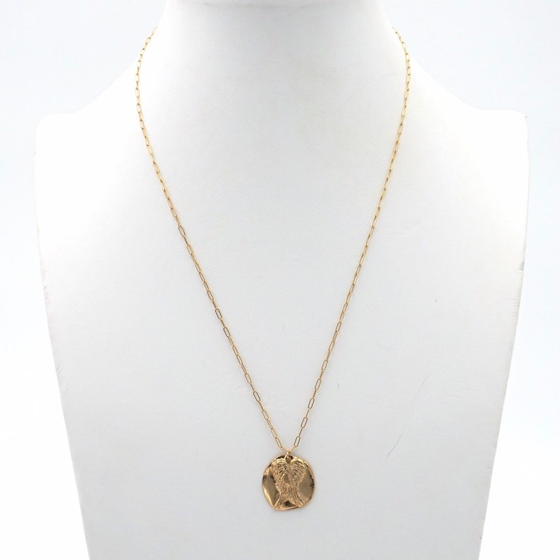 May include: A gold-colored necklace with a pendant featuring a detailed design. The pendant is circular and has an intricate pattern. The necklace is displayed on a white mannequin.