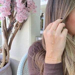 May include: A gold chain bracelet with a delicate design. The bracelet is worn on the wrist and has a long chain that extends up the arm to the finger. The chain is made of small, square-shaped links.