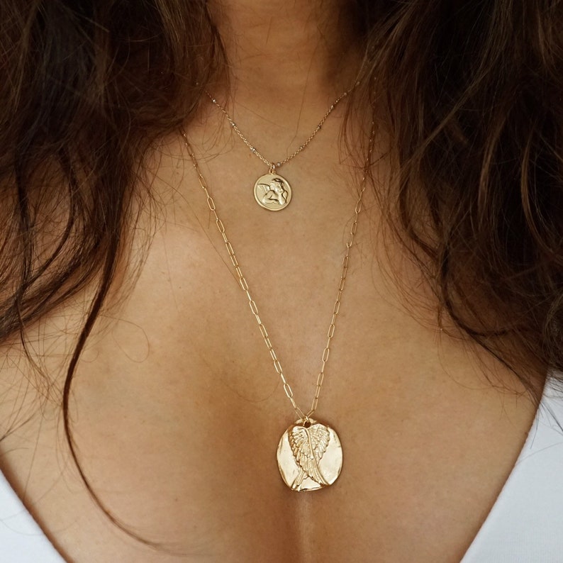 May include: Two gold necklaces. The top necklace has a small round pendant with a figure. The bottom necklace has a larger, oval pendant with an angel wing design. Both necklaces are on a delicate chain.