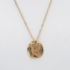 May include: A gold-colored necklace with a pendant featuring an embossed design of angel wings. The pendant is circular and has a textured surface. The chain is a delicate link style.