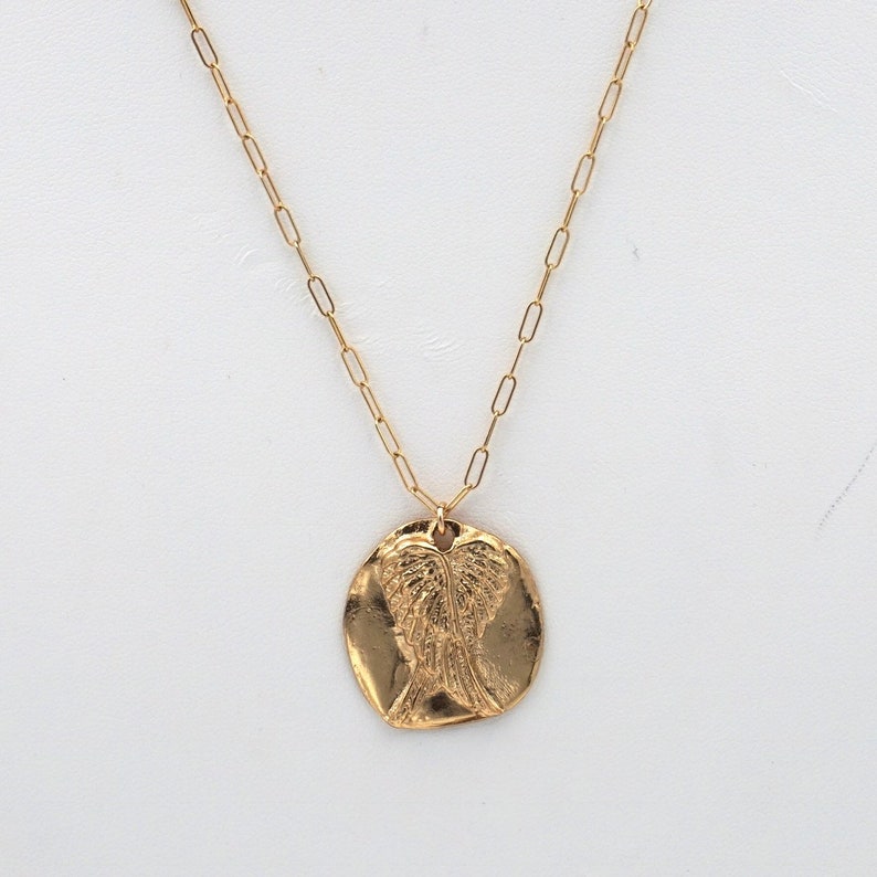 May include: A gold-colored necklace with a pendant featuring an embossed design of angel wings. The pendant is circular and has a textured surface. The chain is a delicate link style.