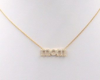 Puede incluir: Un collar de oro con un colgante que deletrea "MOM" en letras de bloque. Las letras están delineadas con pequeñas piedras transparentes.