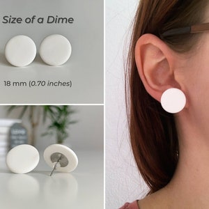 Op de afbeelding: Een paar witte ronde oorbel studs, elk met een diameter van 18 mm (0,70 inch). De oorbellen worden getoond op een wit oppervlak en op het oor van een persoon.