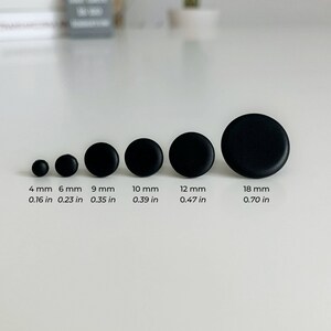 May include: A set of six black round buttons in various sizes, ranging from 4 mm to 18 mm. The buttons are arranged in order of size, with the smallest button on the left and the largest button on the right. The buttons are all made of the same material and have a smooth, matte finish.