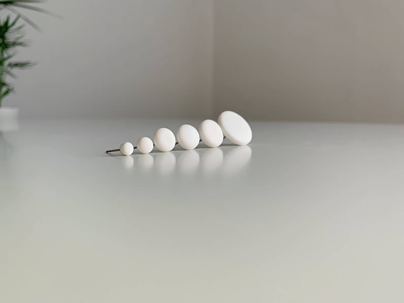 Op de afbeelding: Een set van zeven witte ronde keramische oorbellen in verschillende maten, gerangschikt van klein naar groot.