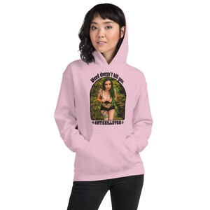 Puede incluir: Sudadera con capucha rosa con un gráfico de una mujer en un fondo verde frondoso. El gráfico dice "Weed doesn't kill you. *KATIEKILLSYOU*"