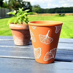 May include: A terracotta flower pot with a white ginkgo leaf pattern. The pot is sitting on a wooden table with a green grassy background.