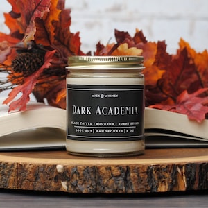May include: A soy wax candle in a glass jar with a black label that reads "Dark Academia." The candle is 8 oz and hand-poured. The background includes fall leaves and an open book, suggesting a cozy, autumnal theme.