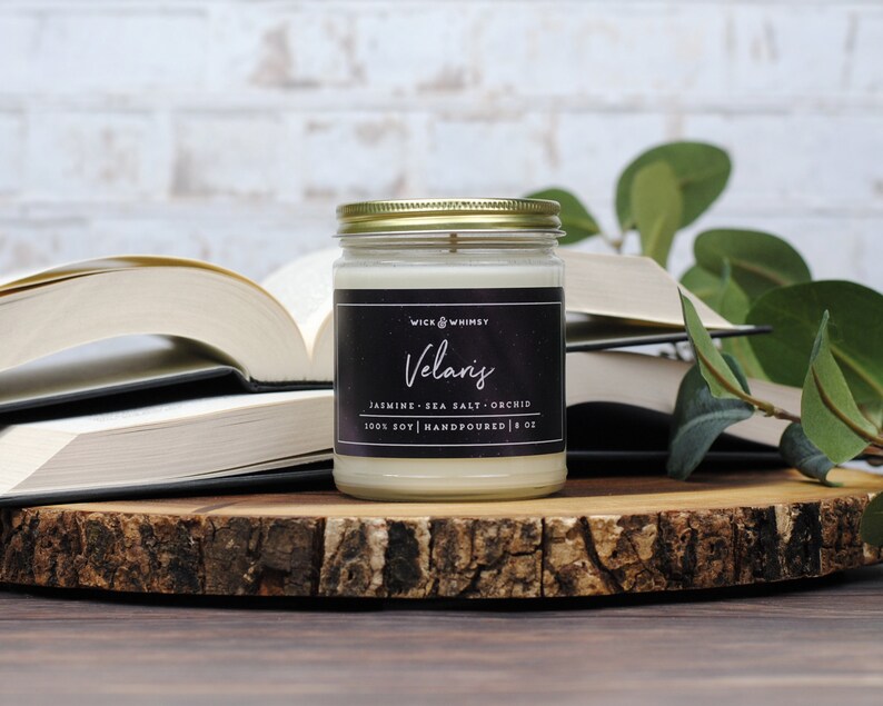 Puede incluir: Una vela de cera color blanco hueso en un frasco de vidrio transparente con tapa dorada y una etiqueta negra con el texto Wick & Whimsy Velaris Jasmine Sea Salt Orchid 100% Soy Handpoured 8 oz, situada frente a unos libros abiertos.