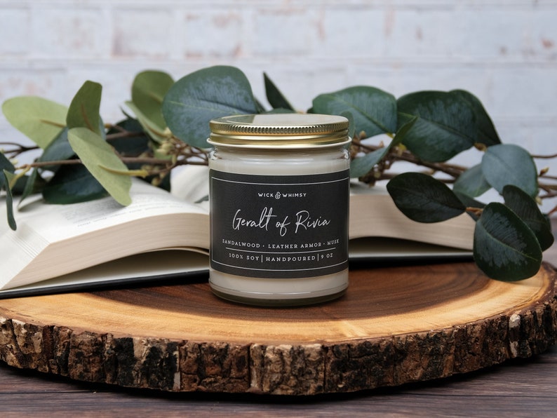 Geralt of Rivia candle, Black label on clear jar with gold lid, sitting on a wood slice with open book in background and eucalyptus branch