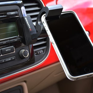 May include: A black Gravity X phone mount attached to a car's air vent, holding a black smartphone with a white case. The phone is positioned horizontally and the mount has a spring-loaded arm.