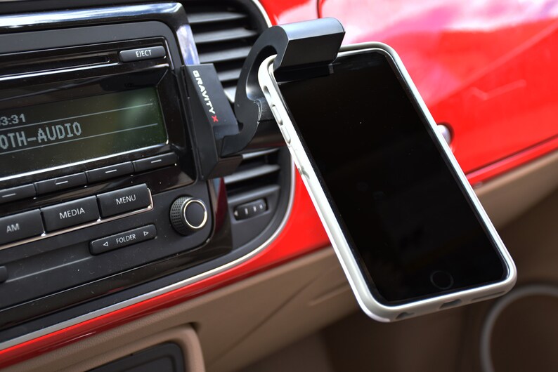 May include: A black Gravity X phone mount attached to a car's air vent, holding a black smartphone with a white case. The phone is positioned horizontally and the mount has a spring-loaded arm.