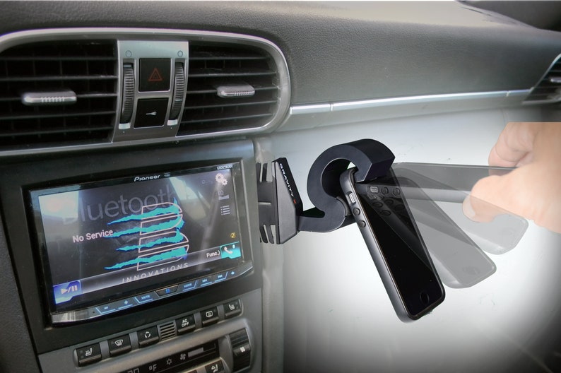 May include: A black car phone mount with a spring-loaded arm to hold a smartphone. The mount is attached to the air vent of the car. The phone is in a black case and the screen is showing the word "Bluetooth" and "No Service".