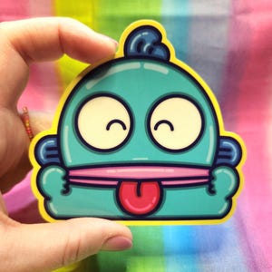 May include: A blue and yellow sticker with a cartoon character with a big smile and a tongue sticking out. The character has large eyes and a blue topknot.