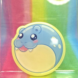 May include: A round, cartoon-style sticker featuring a blue hippo-like character with large, expressive eyes and a smiling mouth. The hippo has pink cheeks and is outlined in yellow. The background is a rainbow of colors.