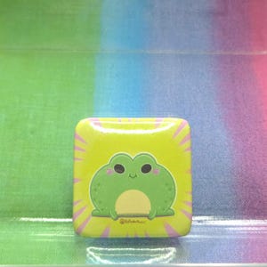 May include: Square button with a cartoon frog design. The button features a light green frog with a yellow belly and black eyes on a yellow background with pink rays. The background is a gradient of green, blue, and pink.