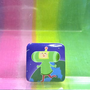 May include: A square pin featuring a green character with a large head and body, running across a blue and green planet. The background is a dark blue with white stars.